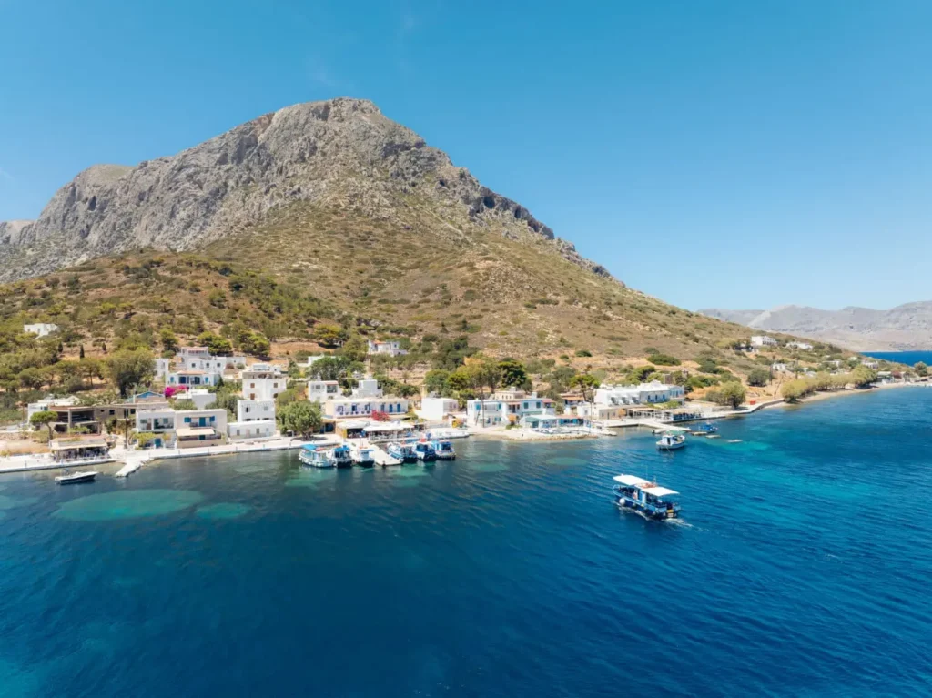 Aerial view of Telendos Island village with waterfront houses, fishing boats, crystal blue sea, and towering mountain backdrop